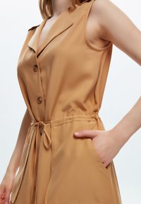 Jumpsuit senza maniche color beige con colletto, chiusura a bottoni frontale, vita elastica e tasche laterali. Realizzato in un tessuto liscio e leggero.