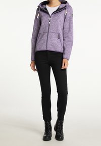 Fialová fleecová bunda se zipem vpředu, kapucí a stahovacími šňůrkami. Má boční kapsy a kontrastní černou podšívku. Kombinována s černými úzkými džíny a botami.
