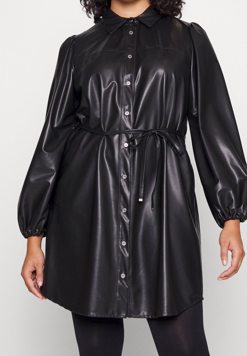 Žena nosící černé koženkové šaty s knoflíky, dlouhými nafouklými rukávy a zavázaným páskem v pase.