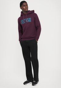 Άνδρας που φοράει μια μοβ �αντιανεμική με μπλε κείμενο, μαύρο παντελόνι με ορατές ραφές, και μαύρα αθλητικά παπούτσια με λευκές σόλες, στέκεται με τα χέρια στις τσέπες.