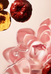 Frutti rossi secchi e gocce d'acqua limpida sono disposti sopra un fiore rosa a fuoco sfocato con petali stratificati su uno sfondo chiaro.