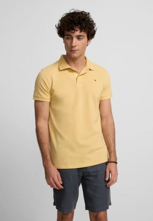 Camisa polo de color amarillo claro, de mangas cortas, con cuello clásico, hecha de algodón, presenta un pequeño logo en el pecho, combinada con pantalones cortos azul marino.