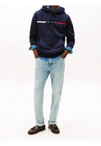 Granatowa bluza z kapturem z białym i czerwonym paskiem, kieszeń z przodu. Jasnoniebieskie jeansy podwinięte przy kostkach. Dopełnieniem stroju są ciemnobrązowe mokasyny.