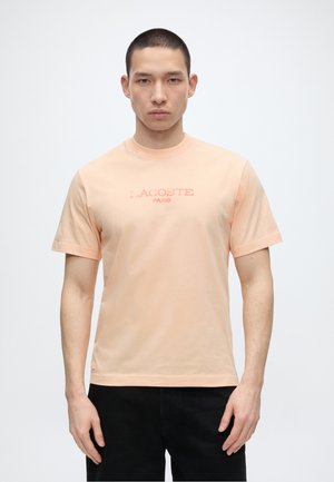 Jeune homme aux cheveux courts porte un t-shirt pêche Lacoste Paris et un pantalon noir, debout devant un fond blanc uni.