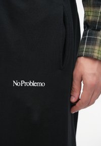 Crne trenirke s bočnim džepom i bijelom izvezenom oznakom "No Problemo" na bedru. Ruka dodiruje tkaninu koja prikazuje mekanu teksturu.