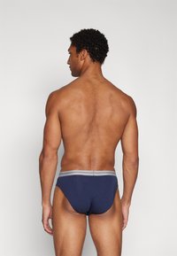 Marine Herren-Briefs mit grauem Bund, aus Baumwolle, eng anliegend und mit minimalistischem Design, von hinten vor einem schlichten Hintergrund gezeigt.