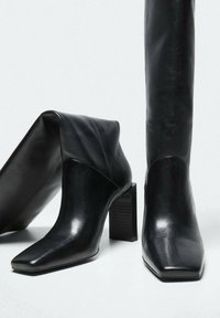 Stivali alla caviglia in pelle nera con punta quadrata, collo arcuato e tacco a blocco testurizzato. Design elegante con texture lisce e lucide.