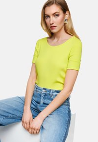 Blonde Frau in einem leuchtend gelben Kurzarmoberteil, hochtaillierten blauen Jeans und großen silbernen Ohrringen, sitzt vor weißem Hintergrund.