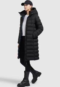 Zwarte pufferjas met capuchon en ritssluiting, voorzien van een gestikte design, zijzakken en een lange snit. Draag het met zwarte leggings en chunky laarzen.