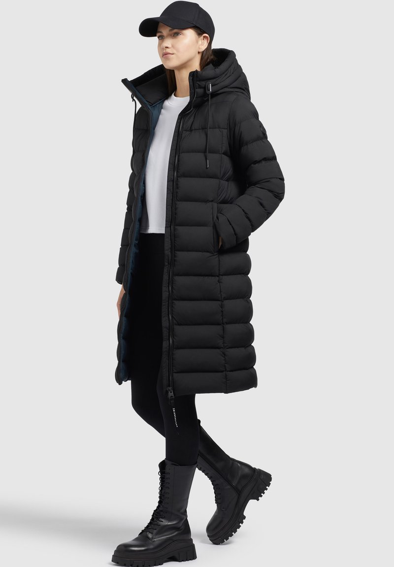 Schwarze Steppjacke mit Kapuze und Reißverschluss, ausgestattet mit einem gesteppten Design, seitlichen Taschen und einem langen Schnitt. Getragen mit schwarzen Leggings und klobigen Stiefeln.