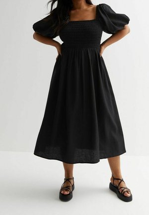 Femme portant une robe midi noire à manches bouffantes et corsage smocké, associée à des sandales plateformes noires, debout les mains sur les hanches.
