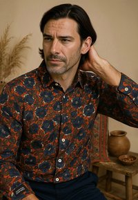 Langarmshirt met een bloemenpatroon van donkere blauwe bloemen op een oranje achtergrond. Het materiaal lijkt textuur te hebben; de knopen zijn licht van kleur.