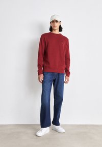 Felpa rossa con scollatura a girocollo e finitura testurizzata, abbinata a jeans blu e sneakers bianche. Il modello indossa un berretto di colore chiaro.