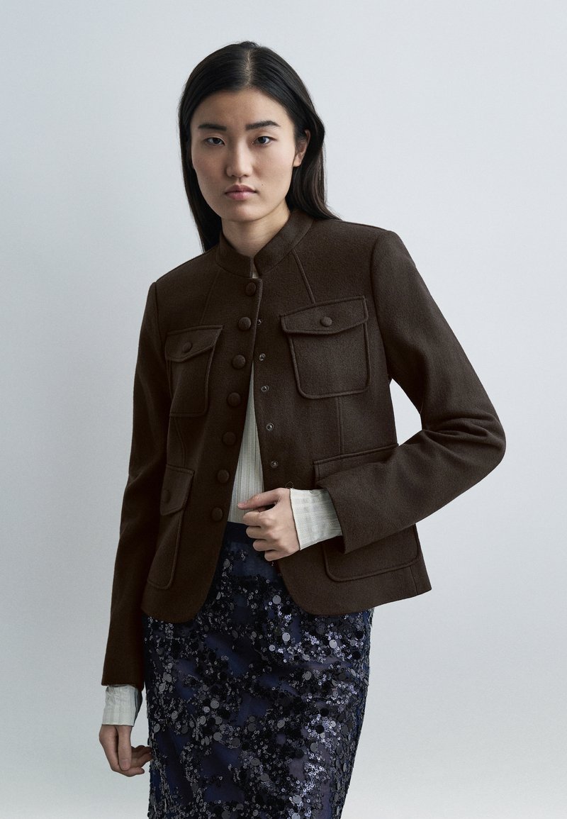 Γυναίκα που φοράει καφέ σακάκι με κουμπιά πάνω α πό λευκό ριπ τοπ και παγιετέ φούστα σε σκούρο μπλε χρώμα, στέκεται μπροστά σε απλό φωτεινό φόντο.