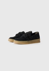 Chaussures en suede noir avec semelle en caoutchouc, dotées de doubles sangles avec boucles et œillets ronds, offrant une silhouette plate et moderne.