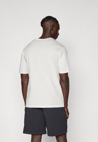 Short-sleeved crew-neck t-shirt in off-white cotton, displayed from the back. Paired with dark shorts, showcasing a simple, clean design.