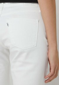 Jeans de denim blanco con detalles en los bolsillos traseros que presentan un patrón de costura curvado. La textura de la tela parece suave y ligeramente texturizada.