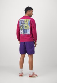 Langærmet maroon skjorte med et farverigt mønster på bagsiden, parret med lilla shorts og pink sko. Hvide sokker synlige; afslappet stil.