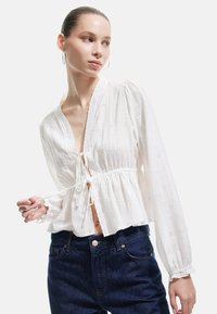 Hvid, tekstureret bluse med frontbinding, løse ærmer, broderi i øjet og en indsnævret talje. Kombineret med mørke denimjeans.