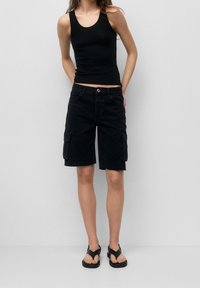 Top negro sin mangas combinado con pantalones cortos de cargo negros que presentan bolsillos laterales, cierre de botón y dobladillo deshilachado. Usado con sandalias negras.