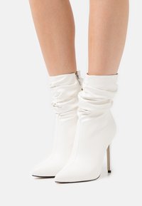 Bottines blanches à design décontracté, bout pointu, haut talon stiletto et fermeture éclair. Fabriquées en matériau synthétique lisse.
