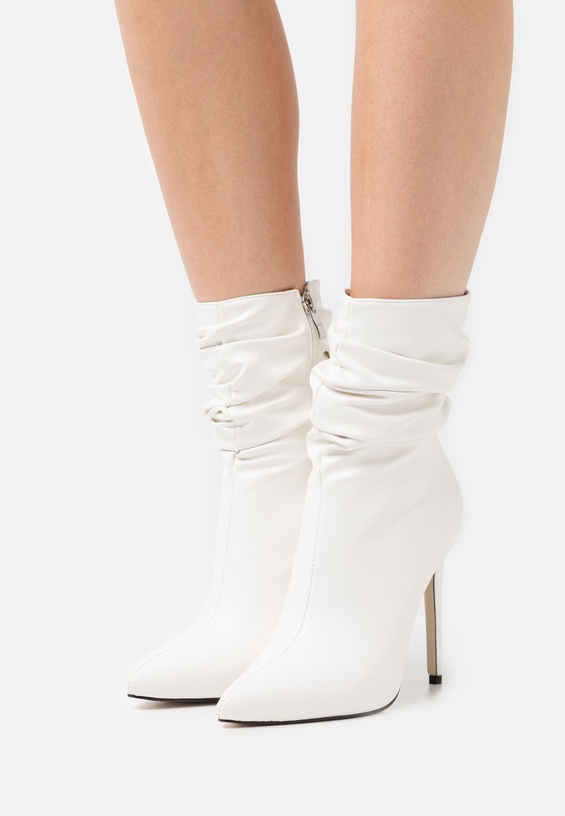 Bottines blanches à design décontracté, bout pointu, haut talon stiletto et fermeture éclair. Fabriquées en matériau synthétique lisse.