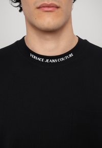 Melns īsā piedurkņu T-krekls ar apaļu kaklu, ar baltu "VERSACE JEANS COUTURE" zīmolu apkaklītē un mazu krūšu kabatu.