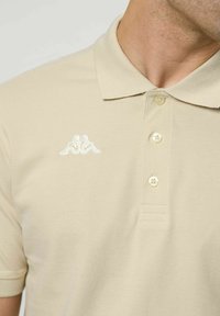 Férfi bézs színű, rövid ujjú galléros pólóban, amelynek háromgombos sliccje van, és a mellkason egy fehér hímzett logó található, amely két ülő alakot ábrázol.