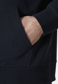 Sudadera azul oscura con un bolsillo lateral, de textura suave, puños de canalé y un ajuste holgado. La tela parece suave y elástica.