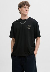 Sort bomulds T-shirt med rund halsudskæring, prydet med et gyldent dekorativt logo på venstre bryst og en afslappet pasform. Glat tekstur.