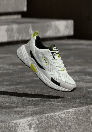 Baskets blanches et noires avec des accents jaunes, en l'air au-dessus du sol en béton et des marches, montrant un mesh respirant et la marque FILA.