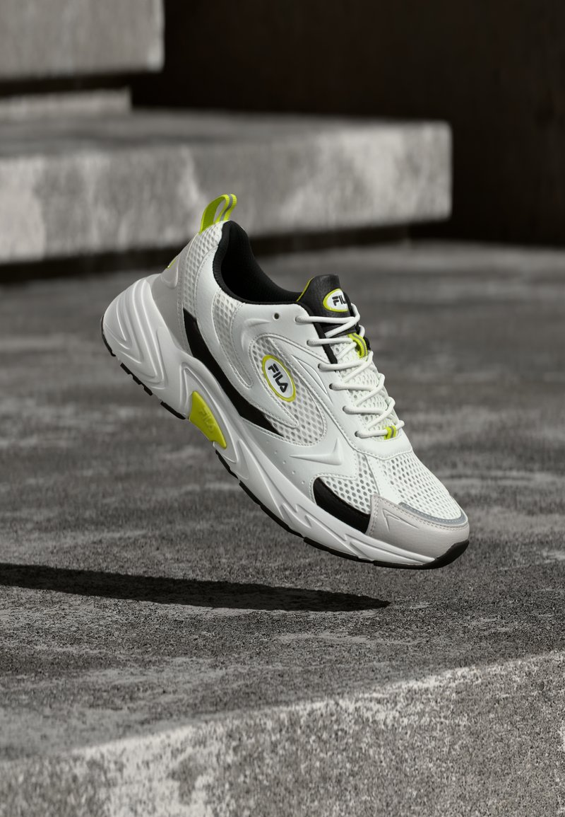 Λευκό και μαύρο αθλητικό παπούτσι με κίτρινες λεπτομέρειες, στον αέρα πάνω από τσιμεντένιο έδαφος και σκαλοπάτια, που δείχνει διαπνέον δίχτυ και τη μάρκα FILA.
