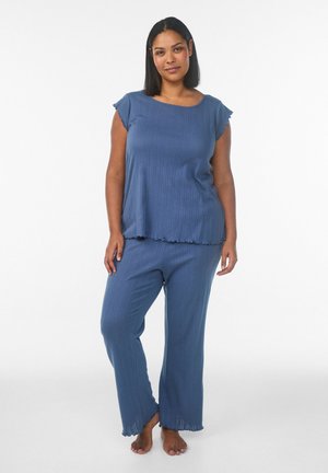 Kvinde står barfodet iført afslappet blå ribstrikket top med korte ærmer og matchende viddeben bukser foran en ensfarvet hvid baggrund.