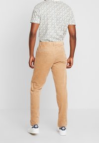 Pantalon en velours côtelé beige avec nervures verticales, coupe structurée et deux poches arrière. Associé à une chemise à manches courtes à motifs et des baskets.