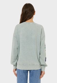 Jasnozielony, oversizowy sweter z prążkowanymi mankietami i dolnym ściągaczem, z delikatną teksturą. Wyróżniające się łatki na rękawach w różnych kolorach.