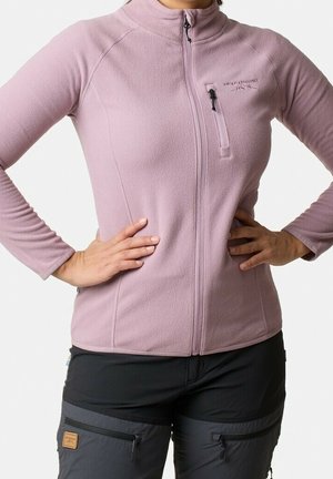 Femme portant une veste polaire ajustée violet clair à fermeture éclair avec une poche poitrine et un pantalon d'extérieur gris foncé avec des poches zippées.