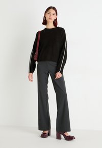 Maglione nero a coste con strisce laterali a contrasto, abbinato a pantaloni a gamba larga grigio scuro e scarpe con tacco a blocco bordeaux. Borsa a spalla rossa.