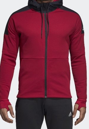 Mand iført en rød hættetrøje med lynlås med sorte skuldre og hætte, samt sorte bukser med Adidas-logo på venstre lår.