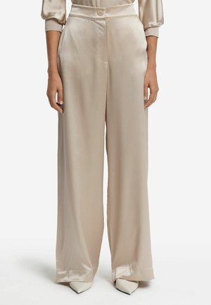 Beige satinbukser med vide ben, frontknap og sidelommer, båret med cremefarvede spidse hæle, vist fra talje til fødder.