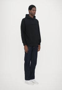 Black hooded sweatshirt with a front pocket, featuring embossed text. Paired with dark denim jeans and white sneakers.