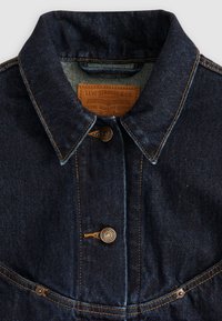 Tamno traper jakna s klasičnim šiljastim ovratnikom, kopčama u bakrenoj nijansi i kontrastnim narančastim šavovima. Kožni brend oznaka unutar ovratnika.