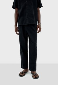 Chemise noire à manches courtes, texturée, et pantalon ample assorti avec une coupe décontractée, associés à des tongs noires sur des pieds nus.