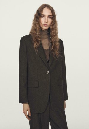 Persona con lunghi capelli mossi, che indossa un blazer oversize marrone scuro e pantaloni coordinati sopra un dolcevita trasparente marrone, su uno sfondo neutro.