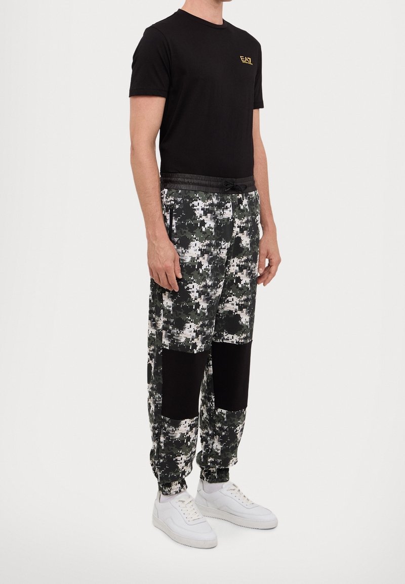 Svart t-shirt med korta ärmar och guldlogga, matchad med joggers i kamouflagemönster i grönt, svart och vitt. Vita sneakers fullbordar outfiten.