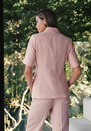 Kobieta ubrana w blado-różową marynarkę z krótkim rękawem i dopasowane spodnie stoi w pomieszczeniu tyłem do widza, przez okno widać zielone drzewa.