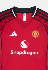 Maglia da calcio rossa con colletto nero e spalle a strisce. Presenta il logo del Manchester United e l'emblema bianco di Snapdragon sul davanti.