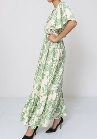 Robe maxi en tissu crème avec des motifs floraux verts, dotée de manches courtes, d'une ceinture à la taille et d'un ourlet à volants. Associée à des talons noirs.