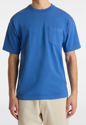 Man draagt een effen blauwe T-shirt met korte mouwen en een borstzak, gecombineerd met lichtbeige broek, staand voor een witte achtergrond.