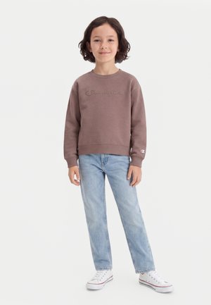 Barn står iført en brun Champion sweatshirt, lyseblå jeans og hvide sneakers mod en ensfarvet lys baggrund.
