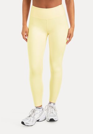 Ben iført lysegule leggings med høj talje, kombineret med hvide sportssko og hvide sokker med sort tekst.